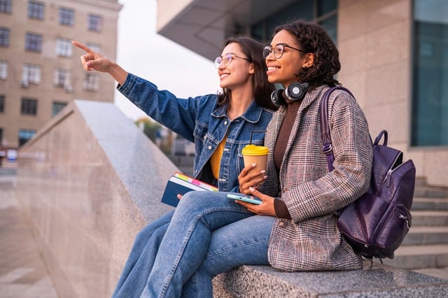 Two smiling college students, one pointing ahead and the other holding a coffee, sit together outdoors.