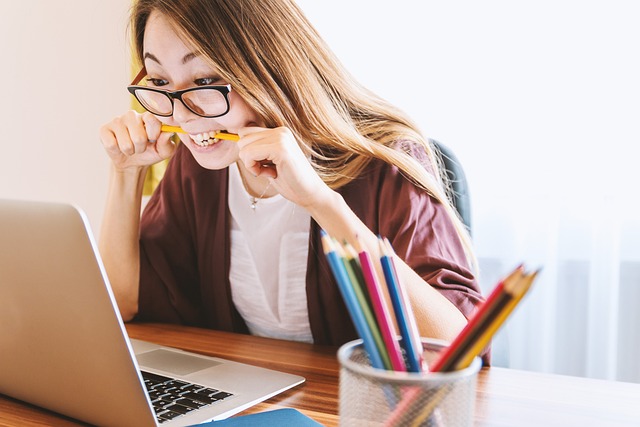 Stressed young woman biting a pencil while working on a laptop, surrounded by colorful pencils in a cup.