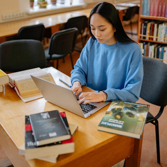 A student working on a laptop surrounded by books, symbolizing research and effort in writing a strong college essay.
